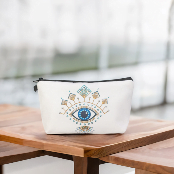The Roula bag, a white cosmetic bag with artistic blue and gold evil eye pattern on a wooden table.