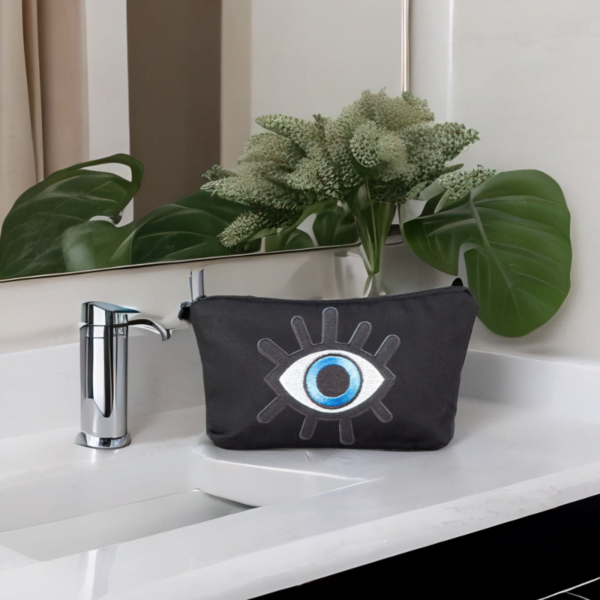 The alia bag, a black cosmetic bag with an artistic blue and black evil eye image in the centre sitting on top of a sink.