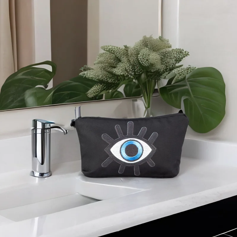 The alia bag, a black cosmetic bag with an artistic blue and black evil eye image in the centre sitting on top of a sink.