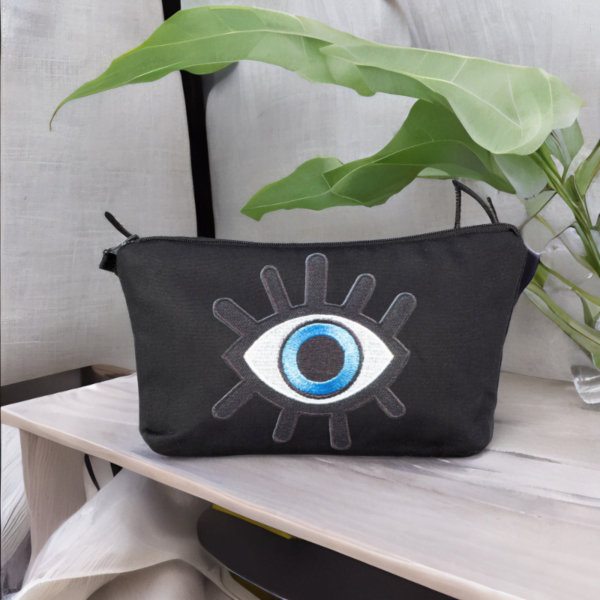 The alia bag, a black cosmetic bag with an artistic blue and black evil eye image in the centre on a wooden surface.
