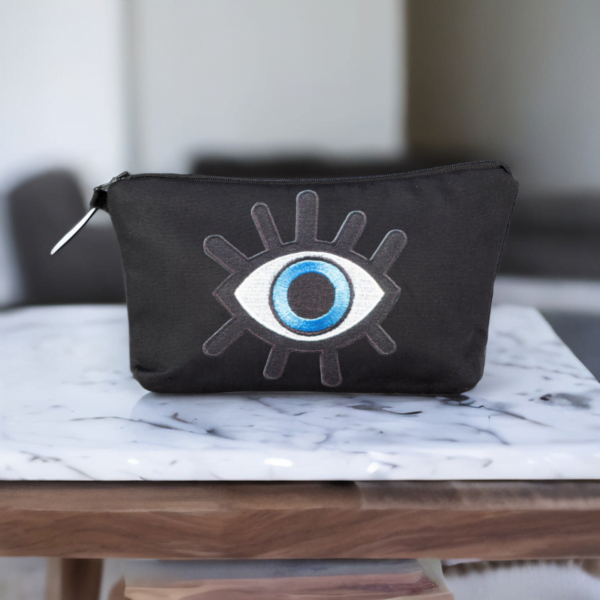 The alia bag, a black cosmetic bag with an artistic blue and black evil eye image in the centre on a small marble table.