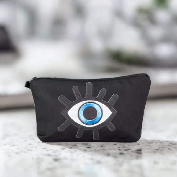 The alia bag, a black cosmetic bag with an artistic blue and black evil eye image in the centre on a marble table.