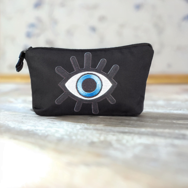 The alia bag, a black cosmetic bag with an artistic blue and black evil eye image in the centre on a marble top.