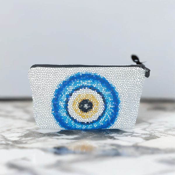 The Nadine bag, a white mosaic patterned cosmetic bag adorned with large blue and yellow evil eye motif in the centre on a marble table.