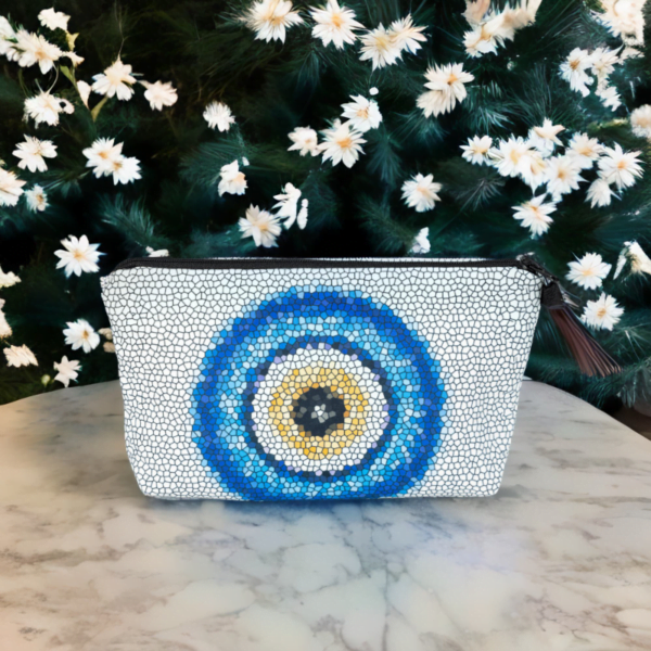 The Nadine bag, a white mosaic patterned cosmetic bag adorned with large blue and yellow evil eye motif in the centre on a table.