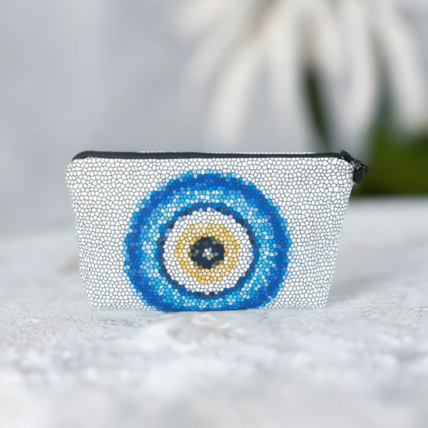 The Nadine bag, a white mosaic patterned cosmetic bag adorned with large blue and yellow evil eye motif in the centre sitting on a marble surface.