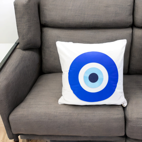 the hiba cushion, a white and plump cushion adorned with artistic blue and white evil eye imagery sitting on a couch.