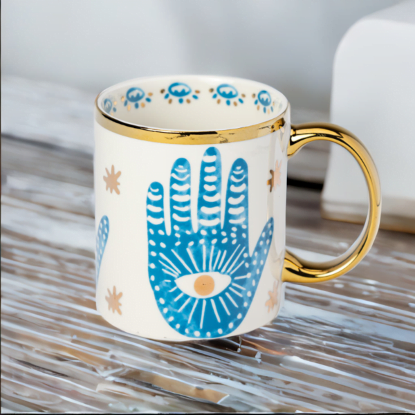A blue and white Hamsa Hand Mug with gold accents on wooden table in London.