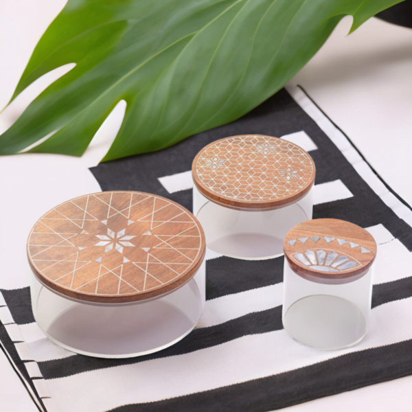 A set of three round storage containers made with transparent acrylic and a wooden lid adorned with quality mother of pearl inlay on a white and black surface.
