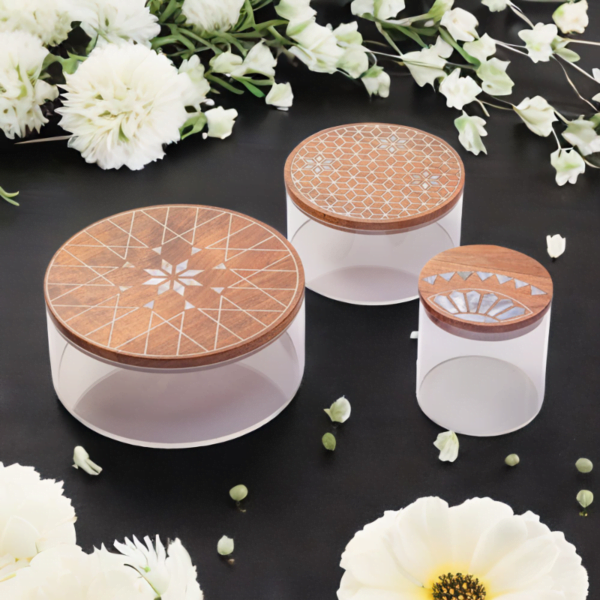 A set of three round storage containers made with transparent acrylic and a wooden lid adorned with quality mother of pearl inlay on a table with flowers on it.