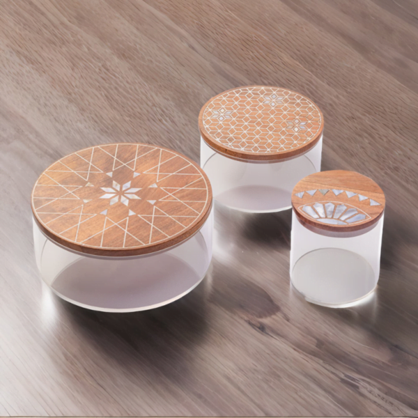A set of three round storage containers made with transparent acrylic and a wooden lid adorned with quality mother of pearl inlay on a wooden table.