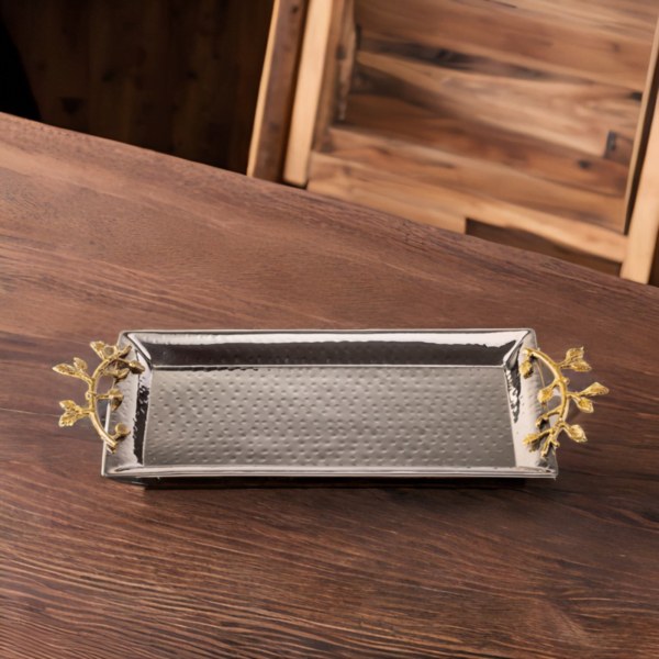 Stainless steel serving tray with hammered styling and gold accented handles sitting on a wooden table.