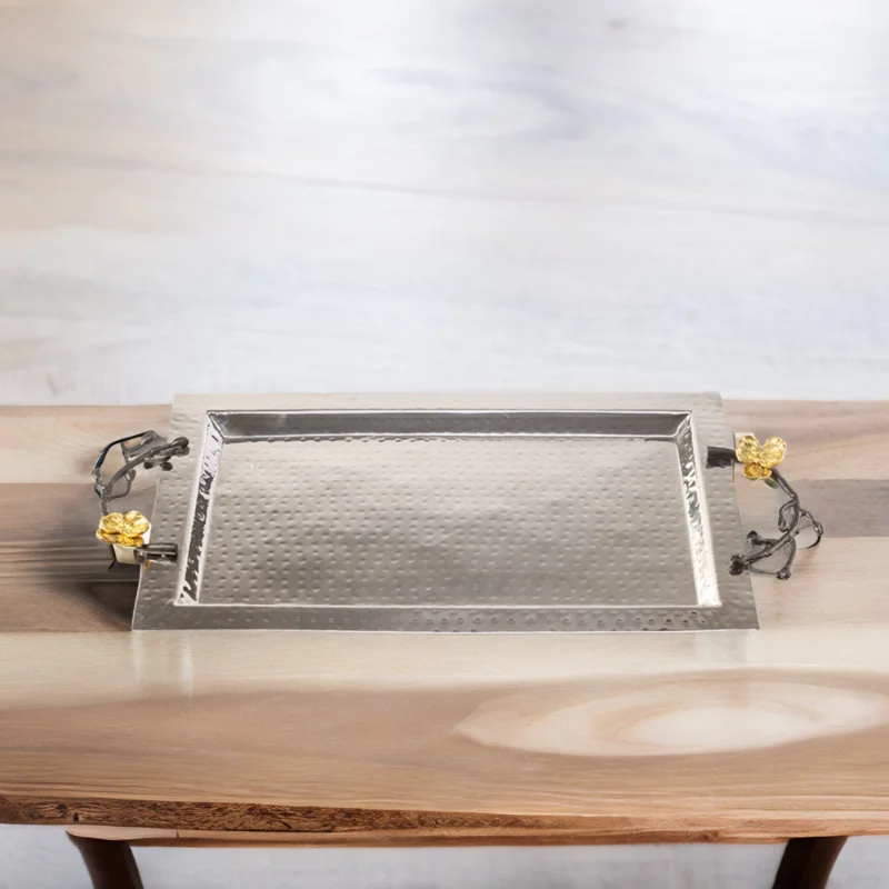 Rectangle hammered stainless steel serving tray with black handles and gold flowers sitting on top of a wooden table.