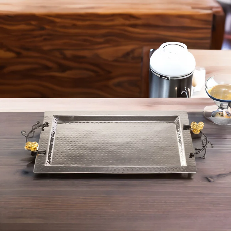 Rectangle hammered stainless steel serving tray with black handles and gold flowers sitting on top of a wooden table.
