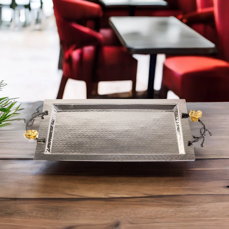 Rectangle hammered stainless steel serving tray with black handles and gold flowers sitting on top of a wooden table.