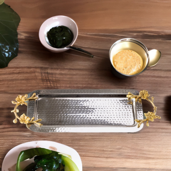 Rectangle hammered stainless steel serving tray adorned with gold handles on a wooden table with plates and cups surrounding it.