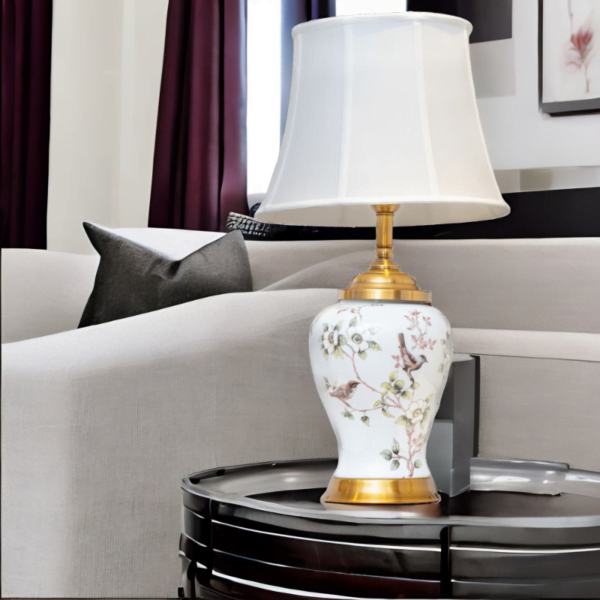 A white porcelain lamp with matching white shade, gold accents and painted flowers and birds on a table in living room.