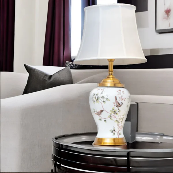 A white porcelain lamp with matching white shade, gold accents and painted flowers and birds on a table in living room.