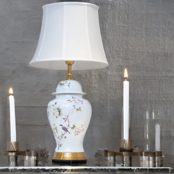 A white lamp with matching white shade, adorned with flowers and bird design, and gold accents on a dining table.