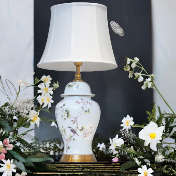A white lamp with matching white shade, adorned with flowers and bird design, and gold accents in a floral surrounding.