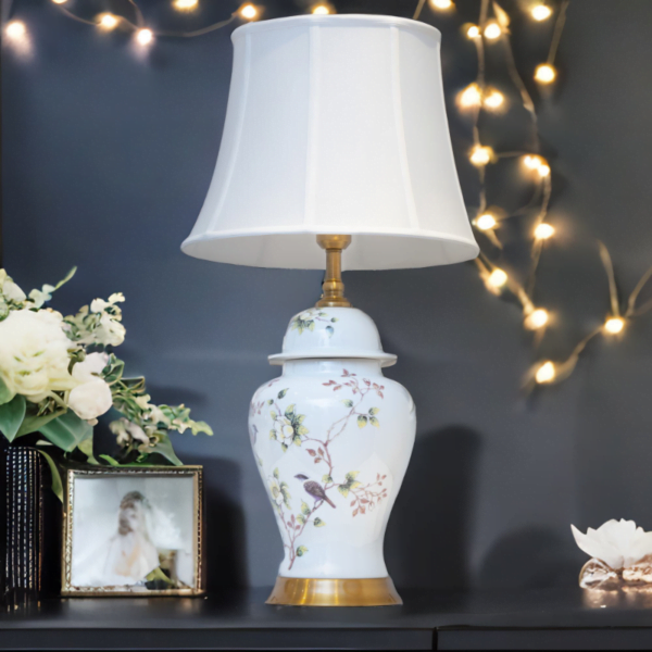 A white lamp with matching white shade, adorned with flowers and bird design, and gold accents on a tv stand.
