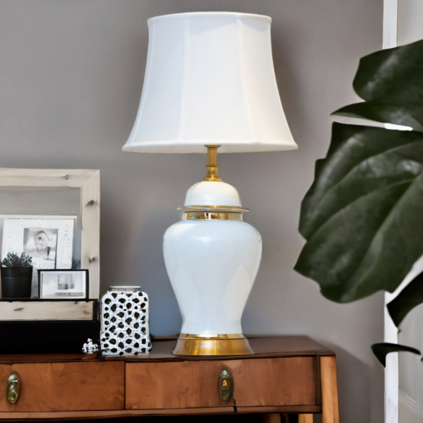 A white lamp with gold accents and matching white shade on a table next to a plant.