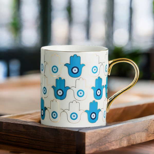 An Azure Hamsa Hand Mug, Blue and White with Gold accents on wooden table.
