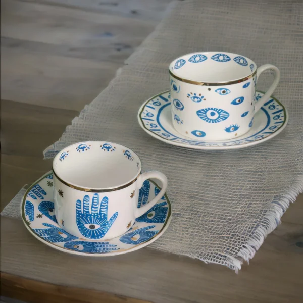 An evil eye and hamsa hand coffee cups with matching saucers on a table.