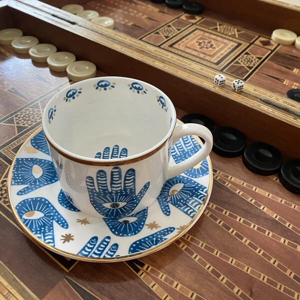 porceline white tea cup with a saucer and hamsa hand prints all with gold edges on a backgammon table.