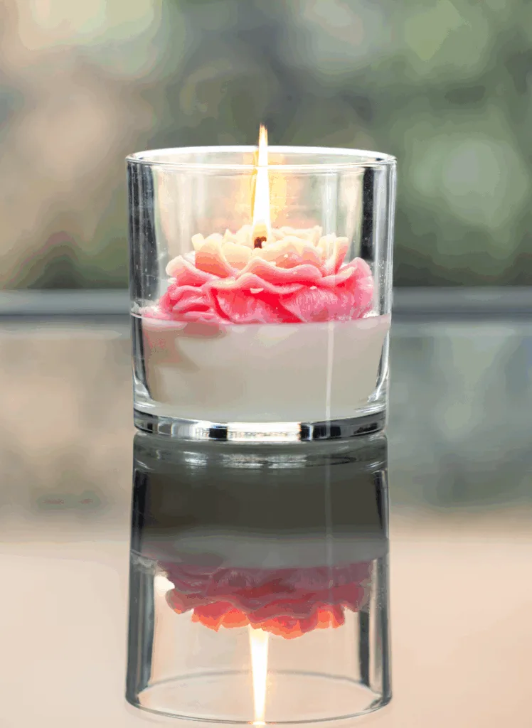 A lit rose candle in a glass jar features a pink and white flower-shaped wax design.
