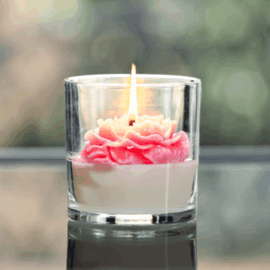 A lit rose candle in a glass jar features a pink and white flower-shaped wax design.