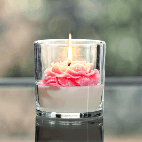 A lit rose candle in a glass jar features a pink and white flower-shaped wax design.