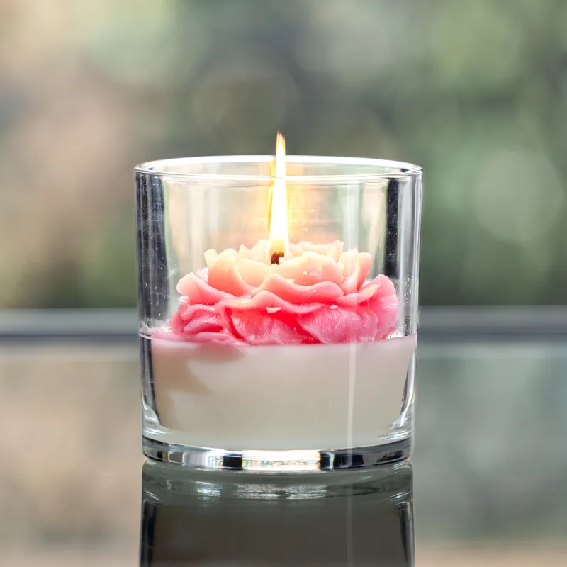 A lit rose candle in a glass jar features a pink and white flower-shaped wax design.