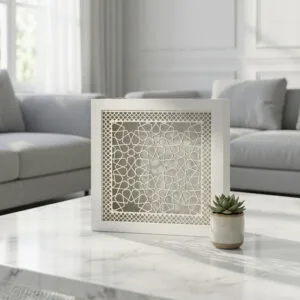 White square decorative box with intricate Islamic geometric arabesque cutout pattern displayed on wooden shelf with fern and books